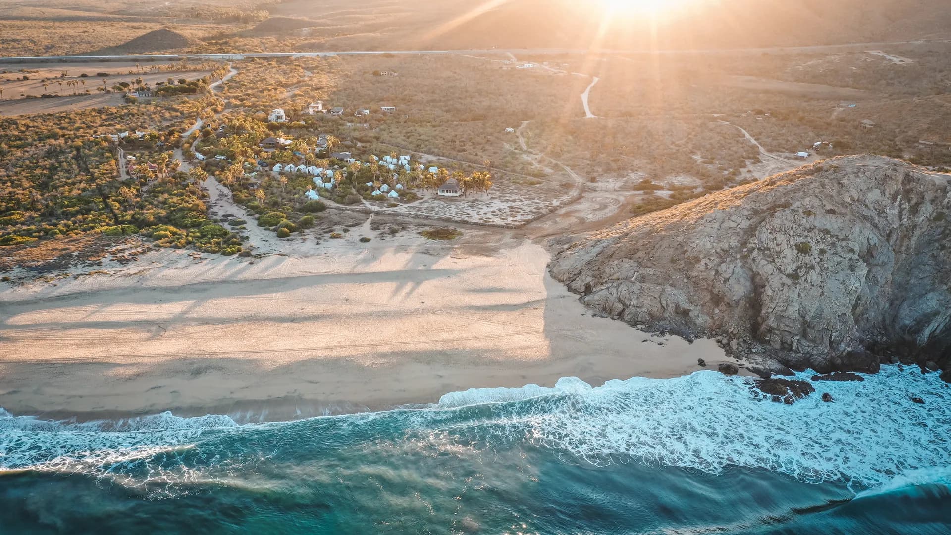 Aerial view of a sunlit Baja beach, rocky headland and surf